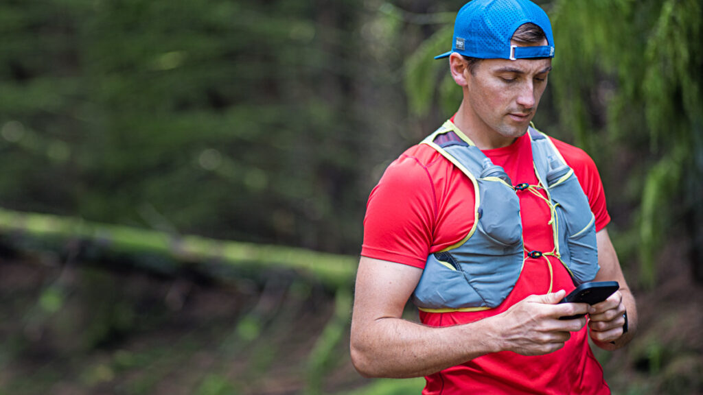 a male trail runner with a running vest on stopped and looking at his smartphone 