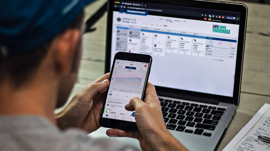 a man looking at the TrainingPeaks app on his phone and laptop in his office