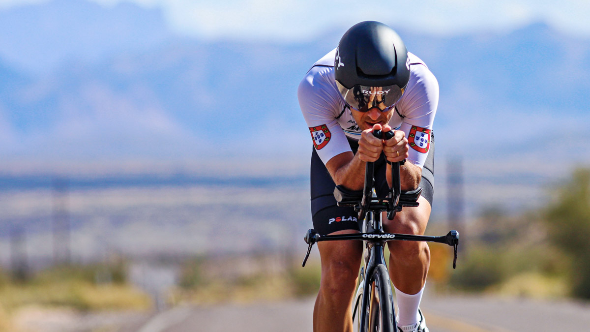 Fit Male Triathlete On Bicycle With Aerobars Rides Down A Road With Mountains In The Background