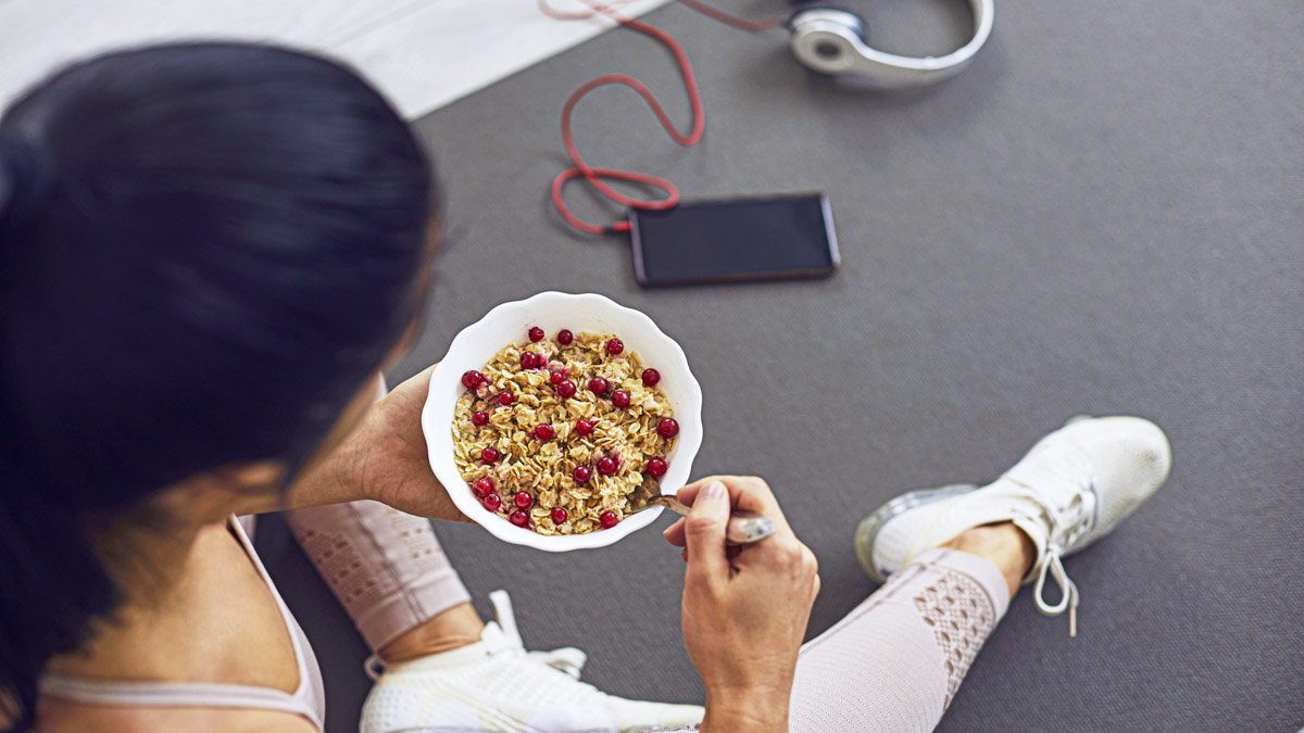 Top View Of A Bowl Of Oatmeal With Cranberries In The Hands Of An Unknown Sports Woman. Woman Sitting On Mat Near Phone And Headphones And Eating Fresh Healthy Food After Active Fitness Workout.