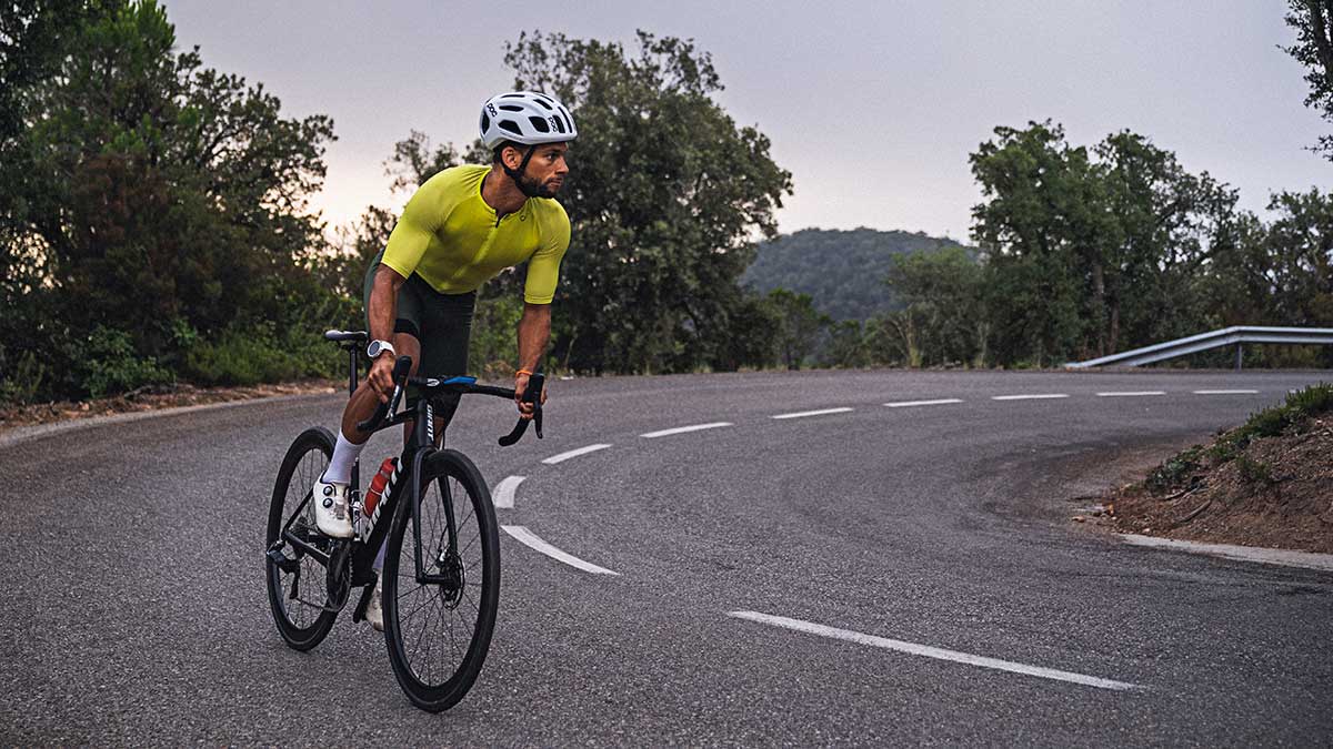 A Male Cyclist Riding Up A Hill On A Paved Road As The Sun Sets Behind Him