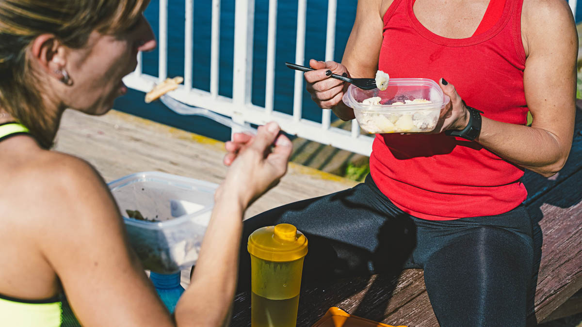 Image Of Two Women Athletes Sitting On The Ground Face To Face Eating Lunch With Healthy Fats