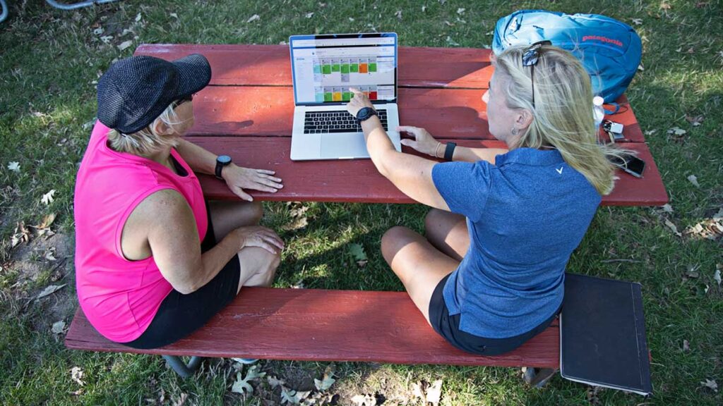 a woman coach sitting with a female athlete on a picnic bench reviewing data on TrainingPeaks