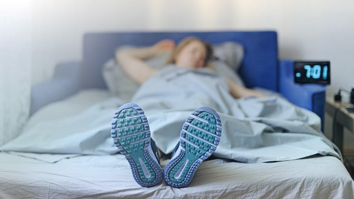 A Woman Laying In Bed Under The Covers With Her Running Shoes On Poking Out From Under The Sheets.