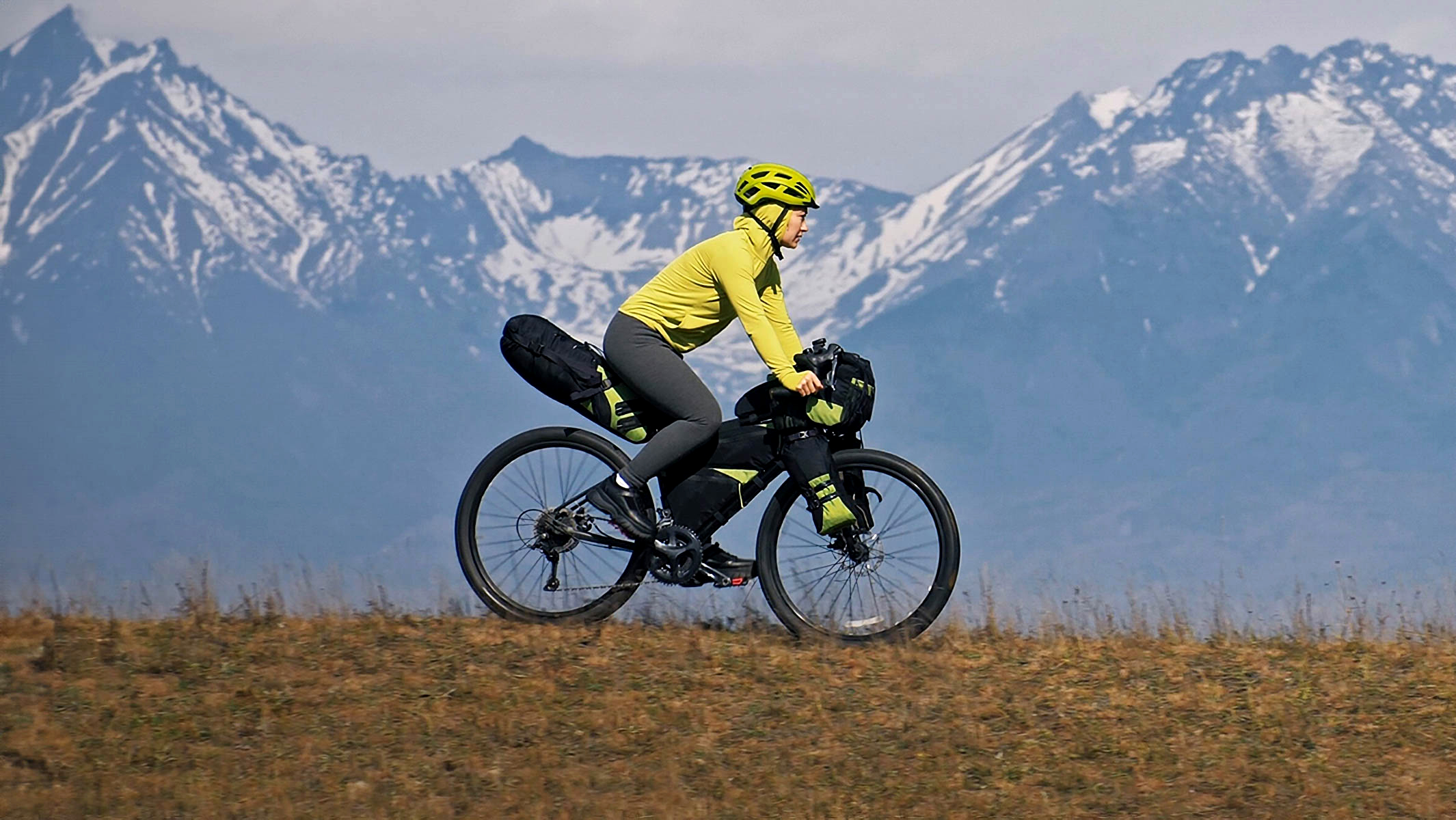 A Woman Traveling On Mixed Terrain Cycle Touring With Bikepacking Bags With A Massive Mountain Range Behind Her..